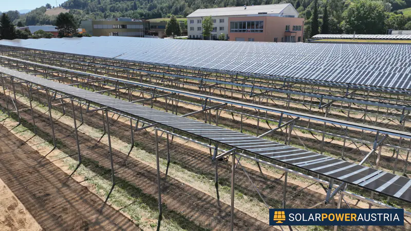 Luftaufnahme einer aufgeständerten Photovoltaik-Anlage über einer Freifläche neben Wohngebäuden